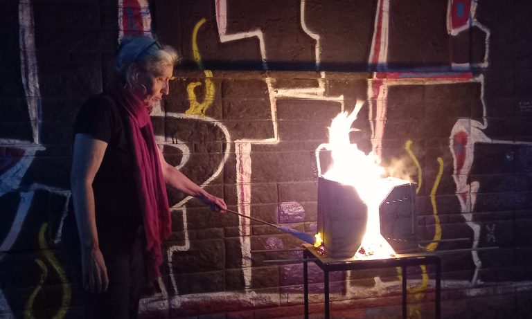 Eine Frau zündet im Dunkeln ein Kartonobjekt auf einem Stahlrohrgestell an. Vor einer Mauer mit Graffity.